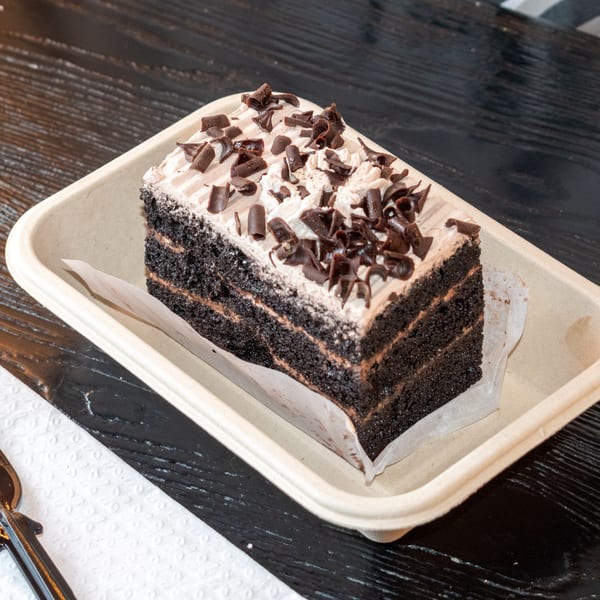 A slice of chocolate cake with cream frosting and chocolate shavings in a disposable container.