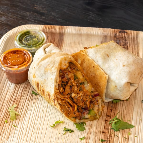 Two Bombay Baking Co. burritos, one cut open to show the filling, with two dipping sauces on a wooden tray.