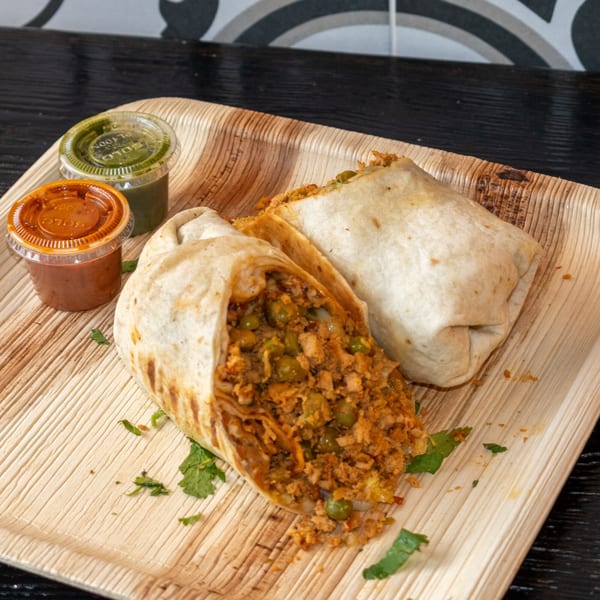 Two Bombay Baking Co. burritos, one cut open, revealing a savory filling with peas, served with two sauces.