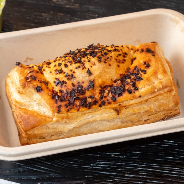 Golden brown Bombay Baking Co. pastry, topped with dark spices and chili flakes, in a beige container.